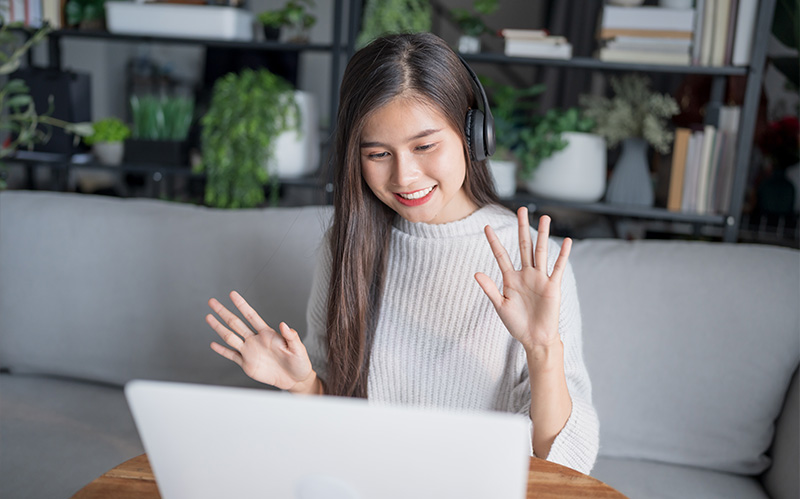 Alt text Smiling woman wearing headphones waving at her laptop during a video call.